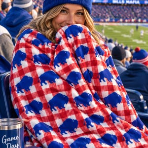 May include: A blue knit beanie with a white pom-pom, the word FOOTBALL, and a football graphic, paired with a red and white checkered fleece blanket featuring a repeating blue buffalo pattern and a blue tumbler with the text Game Day and a heart.