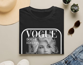 May include: A black t-shirt featuring a white graphic print of a magazine-style cover. The design includes the text VOGUE DOLLY EDITION in white lettering above a black and white portrait of a person with blonde hair, framed by a white border.