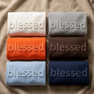 May include: Six folded crewneck sweatshirts arranged in two columns. The colors are white, gray, orange, brown, light blue, and navy blue. Each sweatshirt features the word blessed in raised, matching-colored, three-dimensional lettering.