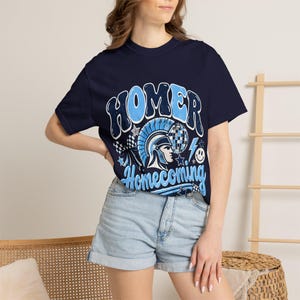 May include: A navy blue short-sleeve t-shirt featuring a graphic print with the text "HOMER" and "Homecoming" in light blue. The design includes a Spartan helmet, a disco ball, a lightning bolt, a smiley face, and checkered patterns.