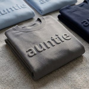 May include: A grey crewneck sweatshirt featuring the word auntie in raised, embossed lettering across the chest. The garment has a ribbed collar and long sleeves, with additional folded sweatshirts in light blue and navy blue visible nearby.