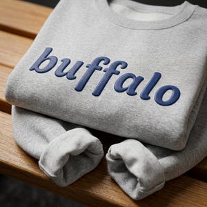 May include: A light gray crewneck sweatshirt featuring the word buffalo in raised, dark navy blue lettering across the chest. The garment has long sleeves with ribbed cuffs and a ribbed neckline, constructed from a soft, heathered fabric.