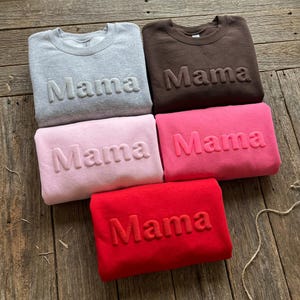 May include: Five crewneck sweatshirts in gray, brown, light pink, dark pink, and red. Each sweatshirt features the word Mama embossed in a matching color across the chest. The garments are arranged in a group, showcasing the textured lettering.