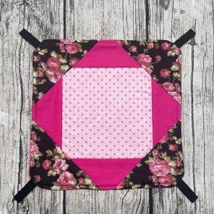 Könnte beinhalten: Ein quadratischer, gesteppter Topflappen mit einem Blumenmuster in Rosa- und Brauntönen. Die Mitte ist hellrosa mit kleinen rosa Punkten. Der Topflappen hat einen leuchtend rosa Rand und schwarze Aufhängeschlaufen an jeder Ecke.