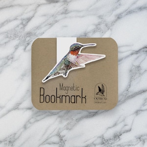 May include: A magnetic bookmark featuring a detailed illustration of a hummingbird with iridescent green, red, and pink feathers. The bookmark is on a brown card with the words "Magnetic Bookmark" and a small logo.