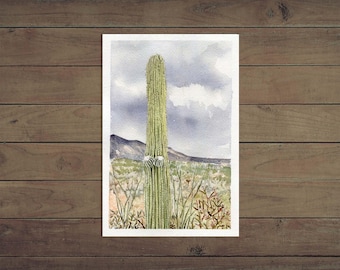 May include: Watercolor painting of a tall, green saguaro cactus with two white flowers. The background features a landscape with mountains, desert vegetation, and a cloudy sky. The artwork is framed in white.