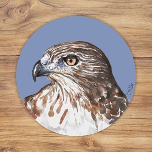 Op de afbeelding: Ronde onderzetter met een aquarel schilderij van een valkenkop in profiel. De valk heeft bruine en witte veren, een zwarte snavel en een gele oog. De achtergrond is een lichtblauwe cirkel en de handtekening van de kunstenaar staat rechtsonder.