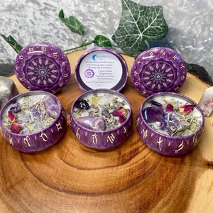 May include: Four purple and gold zodiac candles with amethyst crystals and dried flowers inside. The candles are in small round tins with a mandala design. The text on the label reads "All Natural Soy Candle infused with herbs, oils & gemstones.  Each candle is a powerful tool for spiritual connection, enlightenment & manifestation."