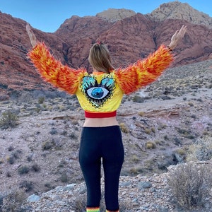 May include: A woman wearing a yellow and orange faux fur jacket with a sequined heart-shaped patch featuring a blue eye. She is standing in a desert landscape with red rock formations in the background.