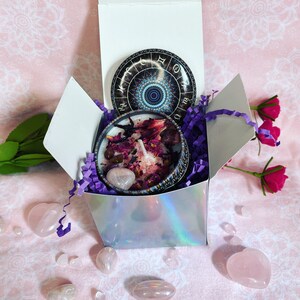 May include: A white gift box with a purple and white shredded paper filling. Inside the box is a candle with a pink and purple design and a rose quartz crystal. The lid of the box has a round sticker with a zodiac wheel design.