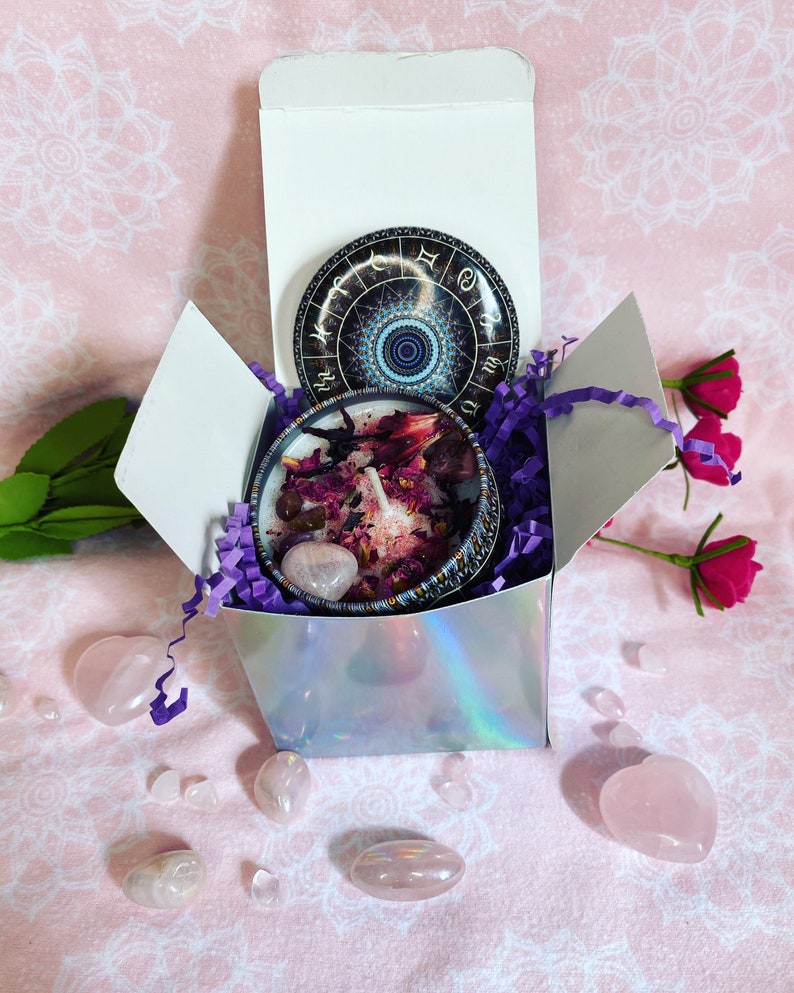 May include: A white gift box with a purple and white shredded paper filling. Inside the box is a candle with a pink and purple design and a rose quartz crystal. The lid of the box has a round sticker with a zodiac wheel design.