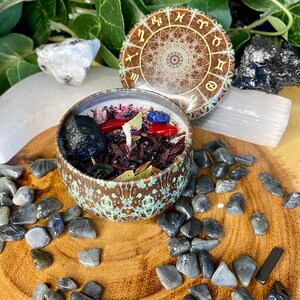 May include: A brown candle jar with a green and gold design. The jar is filled with red, black, and blue crystals and herbs. The lid of the jar has a zodiac wheel design.