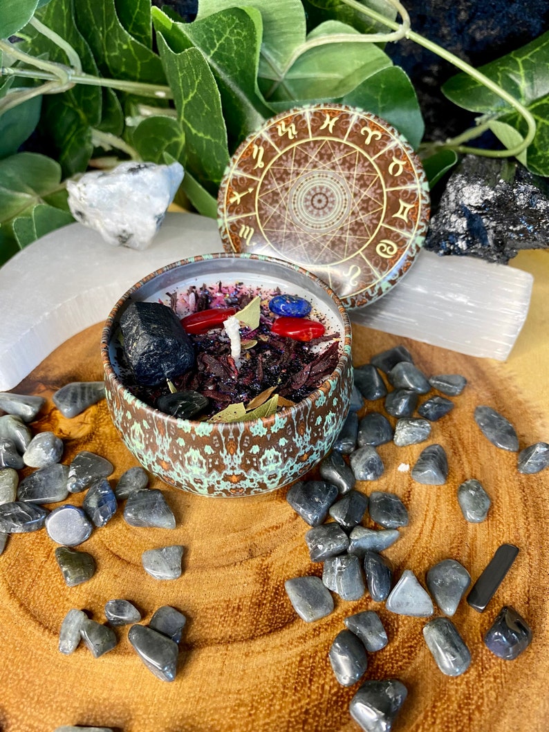 May include: A brown candle jar with a green and gold design. The jar is filled with red, black, and blue crystals and herbs. The lid of the jar has a zodiac wheel design.