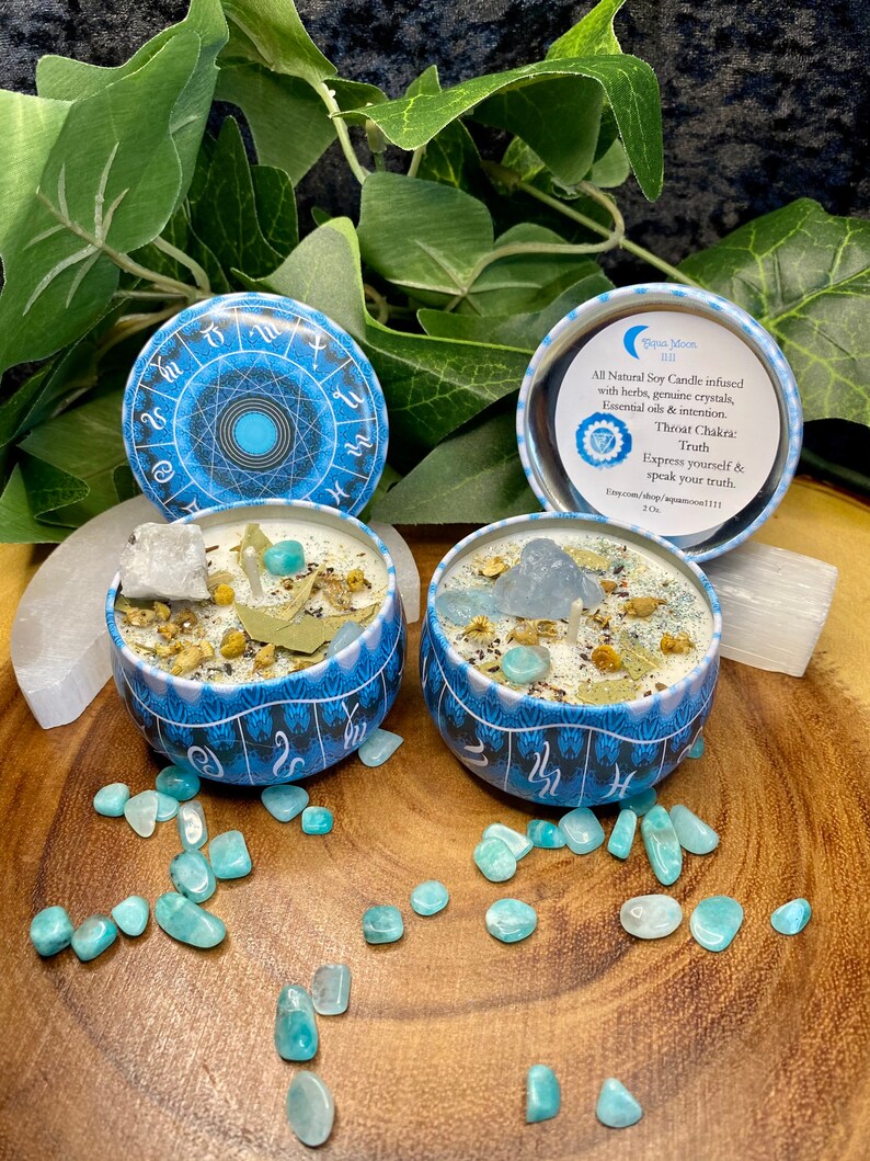 May include: Two blue and white soy candles with a zodiac wheel design. The candles are in small metal tins and are surrounded by green and blue stones. The text on the candle reads "All Natural Soy Candle infused with herbs, genuine crystals, Essential oils & intention. Throat Chakra: Truth Express yourself & speak your truth. Etsy.com/shop/aquamarine111 2 oz"
