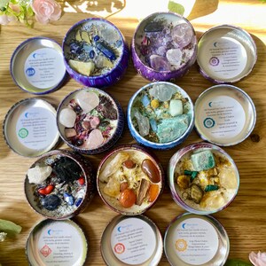 May include: Twelve small, round, metal tins filled with soy wax candles. Each tin has a different color and design, and contains various crystals and herbs. The tins are labeled with the names of the seven chakras, and the text "All Natural Soy Candles infused with herbs & gemstone crystals.  Enhances your energy & intentions."