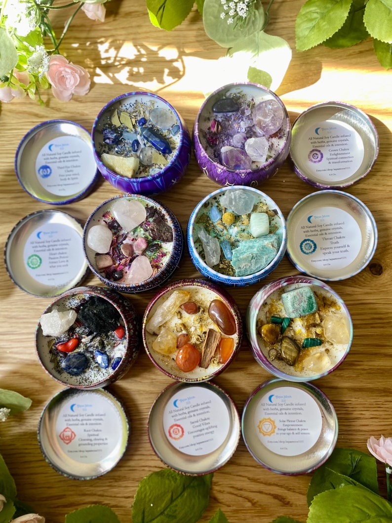 May include: Twelve small, round, metal tins filled with soy wax candles. Each tin has a different color and design, and contains various crystals and herbs. The tins are labeled with the names of the seven chakras, and the text "All Natural Soy Candles infused with herbs & gemstone crystals.  Enhances your energy & intentions."
