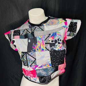May include: A patchwork crop top with a variety of geometric and abstract patterns in black, white, pink, and iridescent colors. The top has short sleeves and a black trim around the neckline and hem.