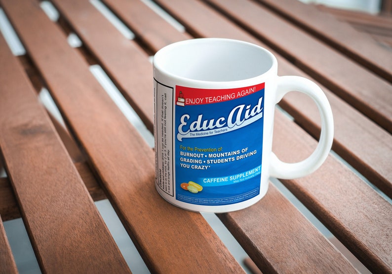 May include: White ceramic mug with a blue and red label that reads "ENJOY TEACHING AGAIN! Educaid The Medicine for Teachers For the Prevention of BURNOUT MOUNTAINS OF GRADING STUDENTS DRIVING YOU CRAZY CAFFEINE SUPPLEMENT"