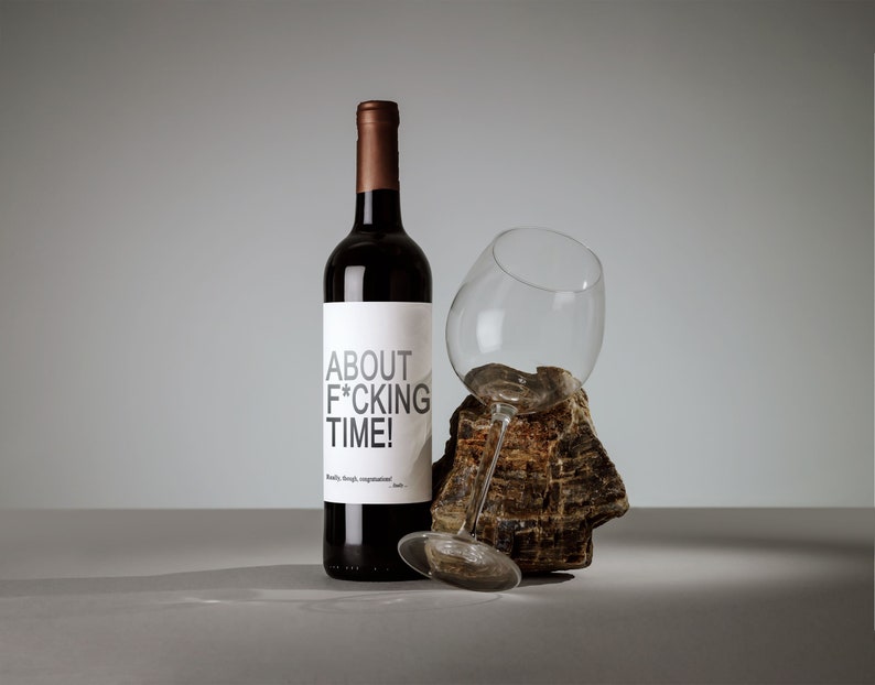 Puede incluir: Una botella de vino tinto con una etiqueta blanca que dice "ABOUT F*CKING TIME!" La botella est&aacute; al lado de una copa de vino sobre una roca.