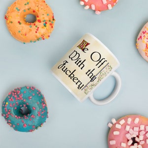 Op de afbeelding: Witte keramische mok met een beige achtergrond en zwarte tekst die "Be Off With thy Juckery" luidt. De mok is omringd door kleurrijke donuts.