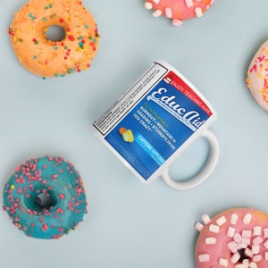 May include: White ceramic mug with a blue and white label that reads "Educ Aid The Medicine for Teachers" and "For the Prevention of Burnout, Mountains of Grading, Students Driving You Crazy. Caffeine Supplement". The mug is surrounded by colourful doughnuts.