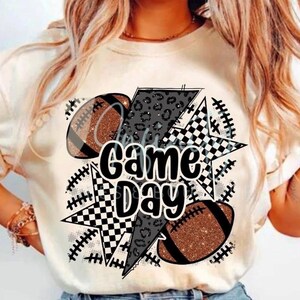May include: Cream-colored t-shirt with a graphic design featuring footballs, stars, and a lightning bolt. The text "Game Day" is prominently displayed in a bold, playful font. The footballs have a glittery texture.