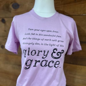 Könnte beinhalten: Helllila T-Shirt mit dem Text "Turn your eyes upon Jesus. Look full in His wonderful face. And the things of earth will grow strangely dim, in the light of His glory & grace." in Schwarz.