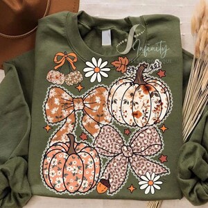 Puede incluir: Sudadera verde oliva con un diseño de temática otoñal. El diseño incluye calabazas, lazos, margaritas y una bellota. Las calabazas y los lazos tienen estampados florales y de leopardo. También se ve el texto "Infinity Tees Boutique".