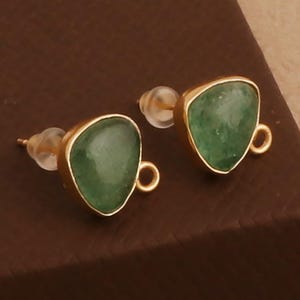 May include: A pair of gold-toned stud earrings with a triangular, green gemstone. The gemstones are set in a gold bezel and have a small loop at the bottom. The earrings are displayed on a brown surface.