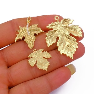 May include: Three gold-colored leaf charms in varying sizes. The largest leaf has detailed veining, while the smallest is a simplified version. One leaf has a line of small, clear stones. All have small loops for hanging.