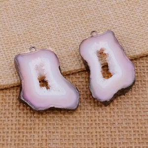 May include: Two polished, irregular-shaped agate pendants. Each pendant features a light purple and white color scheme with a central cavity. The edges are dark gray, and a silver-colored loop is attached for hanging.