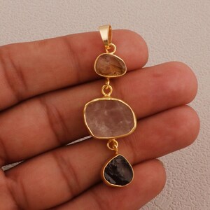 May include: A gold-colored pendant featuring three irregularly shaped gemstones. The top stone is a light brown, the middle is a pale pink, and the bottom is a dark gray-purple. The stones are connected by small gold loops.