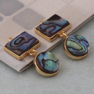 Puede incluir: Un par de pendientes de oro con detalles de concha de abulón azul y verde iridiscente. Los pendientes presentan una pieza rectangular de concha con una pieza redonda más pequeña de concha que cuelga debajo.