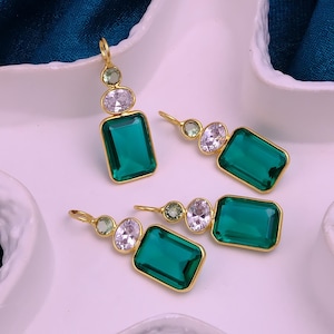 Puede incluir: Tres colgantes dorados con una gran piedra rectangular de color verde esmeralda, una piedra ovalada transparente más pequeña y una diminuta piedra redonda de color verde claro. Cada colgante tiene una pequeña anilla dorada.