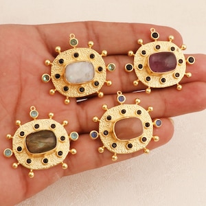 Puede incluir: Cuatro colgantes dorados con piedras rectangulares de diferentes colores. Cada colgante presenta un diseño circular con pequeñas piedras de colores y detalles negros. Las piedras son blancas, moradas, marrones y verdes.