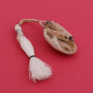 Peut inclure: Un pendentif en pierre naturelle de couleur claire, avec des nuances de marron et de beige, est relié à un pompon blanc par un cordon argenté et doré. Le pompon est fixé avec une bande noire, sur fond rouge.