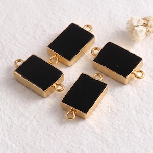 Può includere: Quattro connettori rettangolari per gioielli con un centro nero liscio e solido, incorniciato da un bordo in metallo color oro lucido. Ogni pezzo include due piccoli anelli circolari alle estremità opposte per il fissaggio, disposti sparsi.