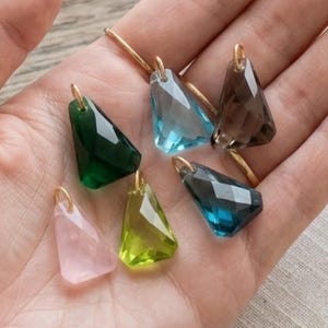 Puede incluir: Un conjunto de seis colgantes de piedras preciosas facetadas con forma trapezoidal en varios colores, incluyendo rosa claro, verde oscuro, verde lima, azul claro, azul oscuro y marrón ahumado, cada uno con una pequeña anilla dorada arriba.