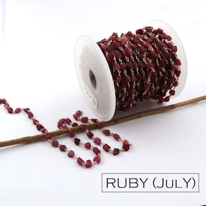 May include: A spool of gold chain with ruby beads. The chain is made of small, irregular ruby beads that are strung together with gold wire. The chain is wound around a white spool with the text "HS-FT" printed on it. The chain is lying on a wooden stick.