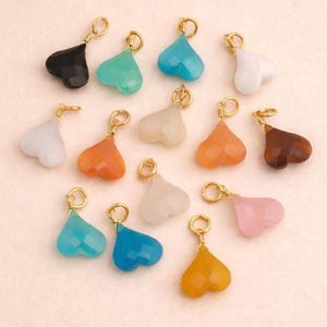 May include: A collection of heart-shaped charms in various colors, including black, teal, blue, white, orange, brown, and pink. Each charm has a gold-colored loop for attaching to jewelry. The charms are faceted and appear to be made of a gemstone or glass.