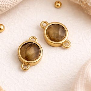 Könnte beinhalten: Zwei runde goldfarbene Schmuckverbinder mit facettierten braunen Steinen in der Mitte. Jeder Verbinder hat zwei kleine Ösen an gegenüberliegenden Seiten zur Befestigung. Drei winzige goldfarbene, kugelförmige Perlen liegen verstreut daneben.