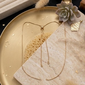 Op de afbeelding: Een verstelbare goudkleurige box-ketting met een schuifkraalmechanisme. De ketting heeft een delicate, dunne schakelstructuur met twee bungelende uiteinden. De ketting wordt gepresenteerd op een lichtgekleurd, getextureerd stenen oppervlak.