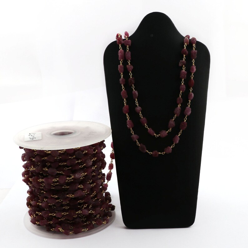 May include: A spool of ruby red gemstone beads strung on a gold chain. The beads are irregular in shape and have a matte finish. The chain is visible between the beads. The spool is white with a black label.