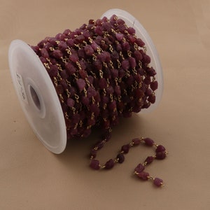 May include: A spool of ruby red gemstone beads strung on a gold chain. The chain is made of small, delicate links. The beads are irregular in shape and have a rough, natural texture.