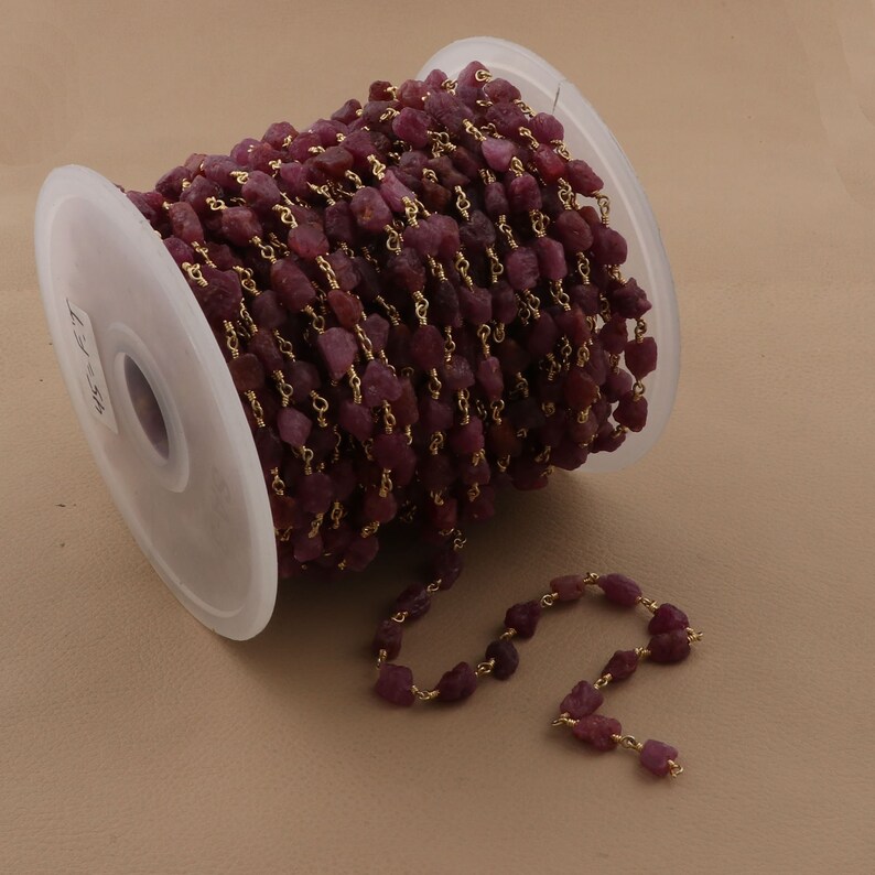 May include: A spool of ruby red gemstone beads strung on a gold chain. The chain is made of small, delicate links. The beads are irregular in shape and have a rough, natural texture.