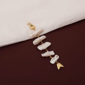 May include: A gold-toned pendant shaped like a fish skeleton, featuring five irregular white baroque pearls as the body segments. The head piece includes a small green stone, and the tail is a textured gold-toned metal fin component.