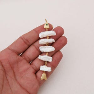 May include: A vertical pendant shaped like a fish skeleton, featuring five elongated white baroque pearls arranged as the body. The head is a gold-toned triangular piece with a small green stone, and the tail is a gold-toned fin-shaped piece.