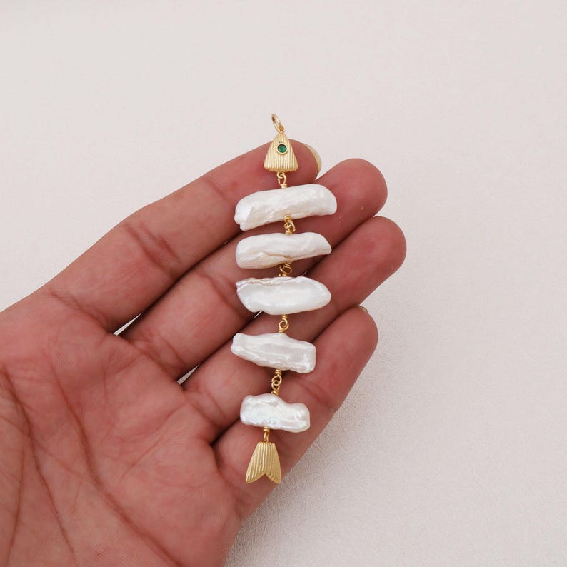 May include: A vertical pendant shaped like a fish skeleton, featuring five elongated white baroque pearls arranged as the body. The head is a gold-toned triangular piece with a small green stone, and the tail is a gold-toned fin-shaped piece.
