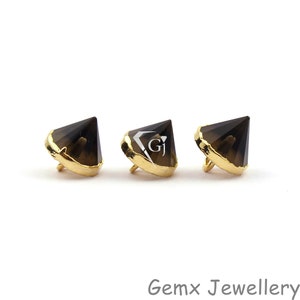Peut inclure: Trois pierres de quartz fumé en forme de pyramide, chacune sertie dans une monture en métal doré avec une boucle sur le dessous. La pierre centrale porte un logo blanc avec les lettres GJ. Le texte Gemx Jewellery est imprimé en gris en bas à droite.