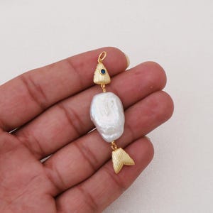 May include: A gold-toned metal pendant shaped like a fish, featuring a textured triangular head with a small blue stone eye, a large irregular white baroque pearl as the body, and a textured gold-toned metal tail piece at the bottom.